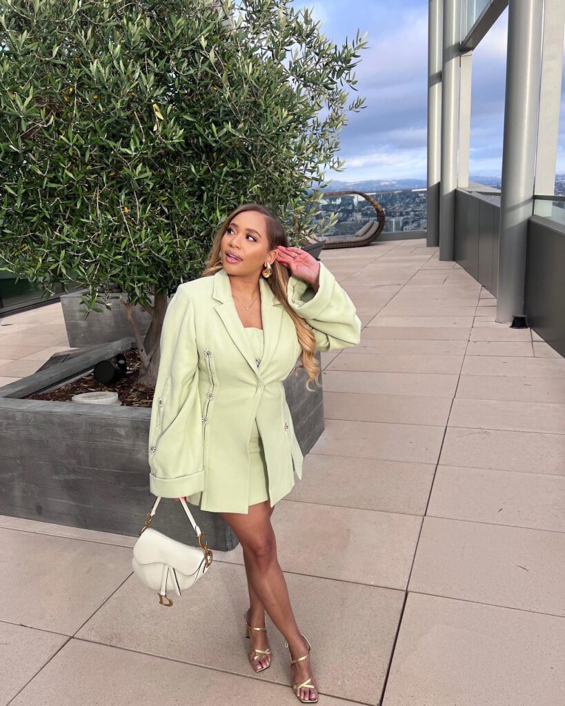 A stylish woman poses on a rooftop patio, wearing a pale green cult gaia blazer and shorts set, gold high heels, and holding a white Dior saddle bag.