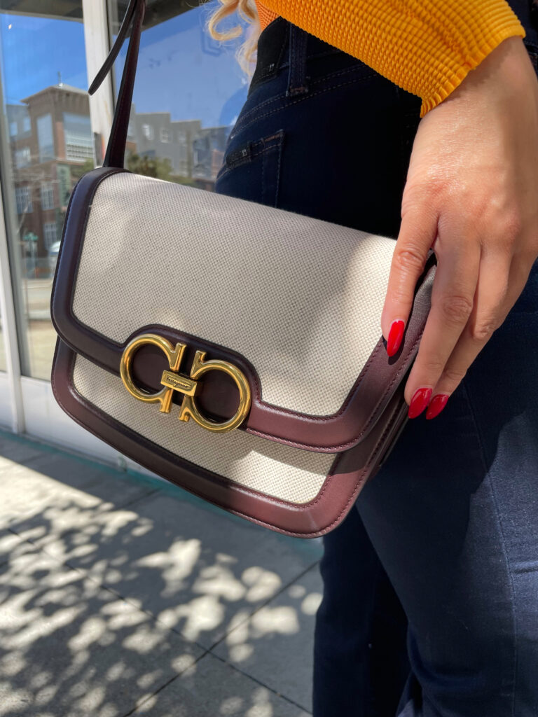 A beige Ferragamo trapezoid-shaped purse on a woman's shoulder, styled with Joe's Jeans petite cut and Jimmy Choo heels